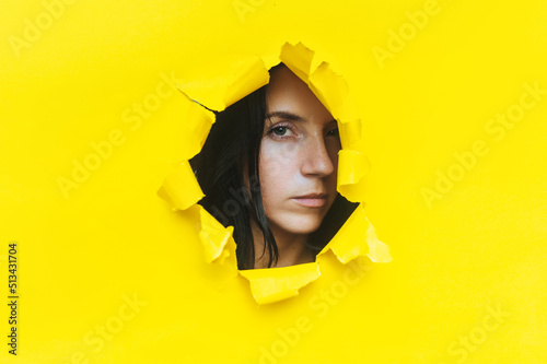 Close-up portrait of a caucasian young woman looking through a hole in yellow paper. An incredulous gaze. Women's curiosity and gossip. Jealous wife. Spy concept. © shchus