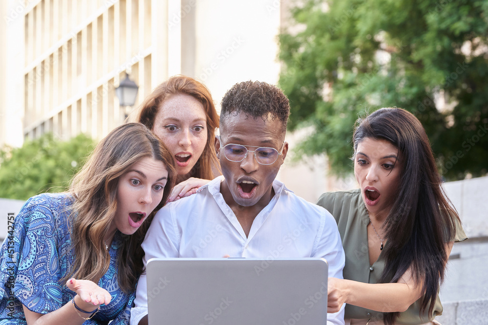 Group of multiracial people looking shocked at the laptop screen while using it outdoors in the street. Urban lifestyle, technology and friendship concept. Group of multiracial people looking shocked at the laptop screen while using it outdoors in the street. Urban lifestyle, technology and friendship concept.