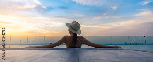 Asian beautiful woman in hat watching the beautiful scenic sunset at the edge of the infinity pool with cityscape, enjoying nature background banner panorama. Woman from behind relaxing at landscape.