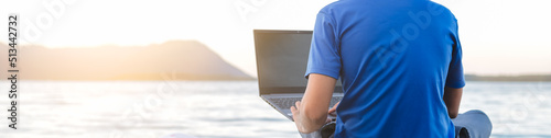 young hipster digital nomad man sitting on wooden pier at sea working on internet remotely at sunset - Traveling with a computer - Online dream job concept - Selective Focus. photo