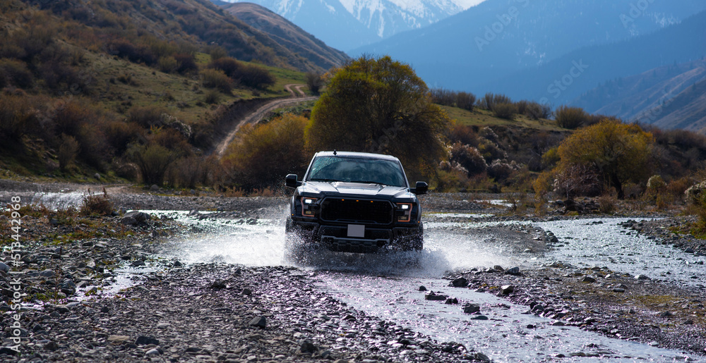Pick-up truck is off road in the small river in the mountains. View with snow covered mountains in the distance. Landscape of country road with beautiful cloud and sky. Beauty of wild nature. Pick-up truck is off road in the small river in the mountains. View with snow covered mountains in the distance. Landscape of country road with beautiful cloud and sky. Beauty of wild nature.