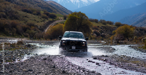 Pick-up truck is off road in the small river in the mountains. View with snow covered mountains in the distance. Landscape of country road with beautiful cloud and sky. Beauty of wild nature.