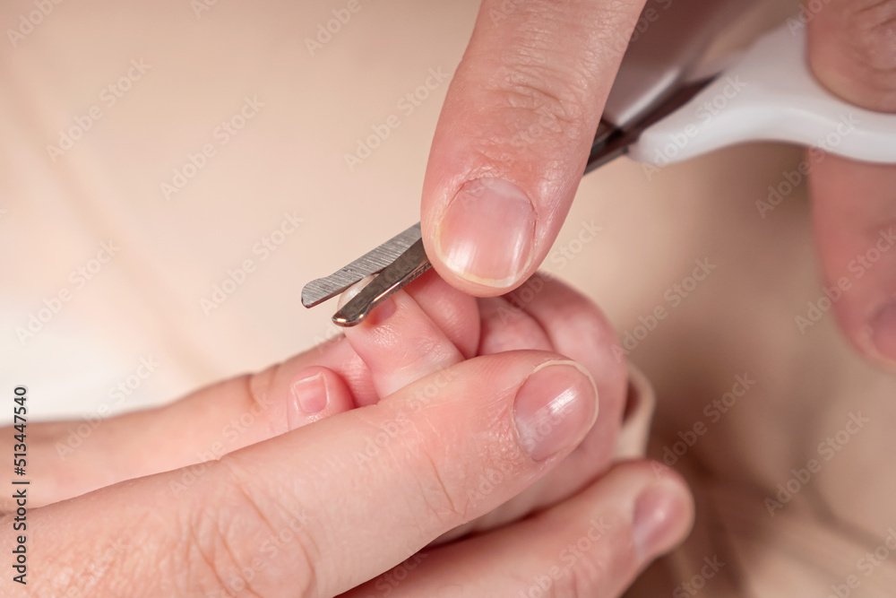 Hands of mother carefully cut tiny nails of baby girl with scissors on blurred background. Woman takes care of hygiene for newborn kid extreme closeup