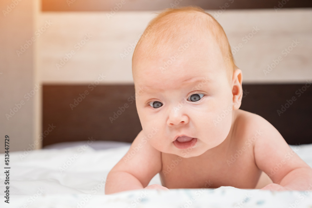 Cute baby girl raises up head lying on bed. Curious newborn kid examines bedroom looking around, sunlight Cute baby girl raises up head lying on bed. Curious newborn kid examines bedroom looking around, sunlight