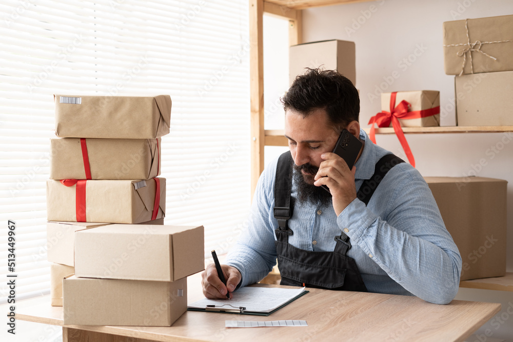 Indian bearded man online seller confirming orders from customer on the phone. E-commerce male business owner using phone in store warehouse. SME