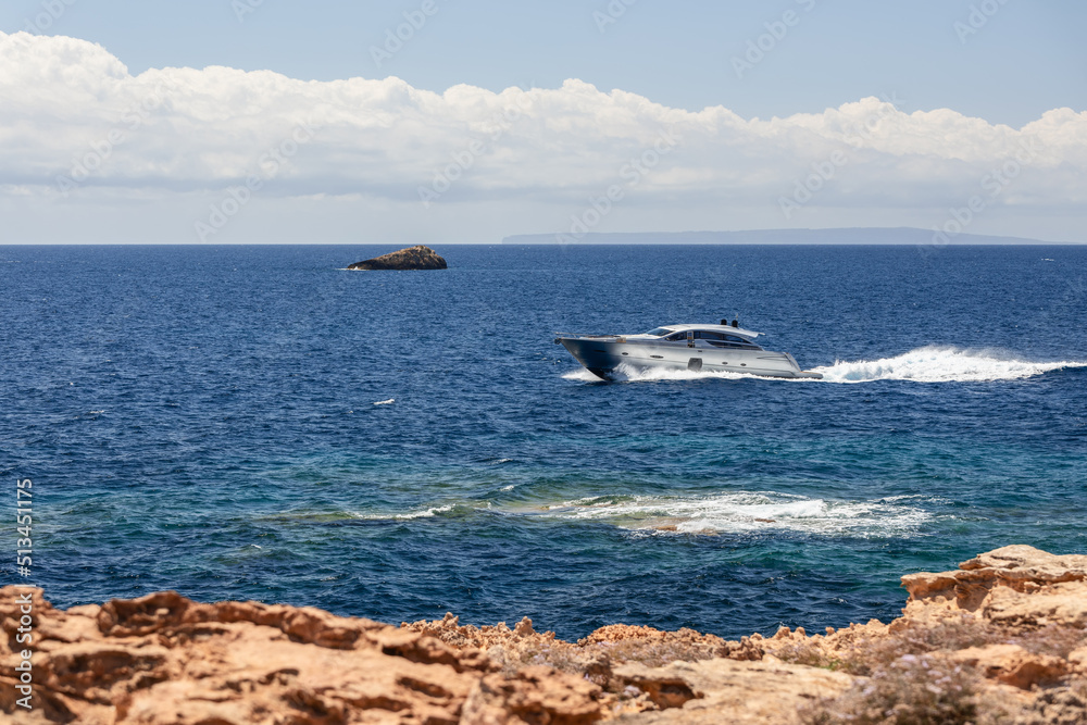 Small silver motor boat moves along the rocky yellow coast of Ibiza island. Clearest calm sea and horizontal line of white solid clouds on the horizon, Balearic Islands, Spain