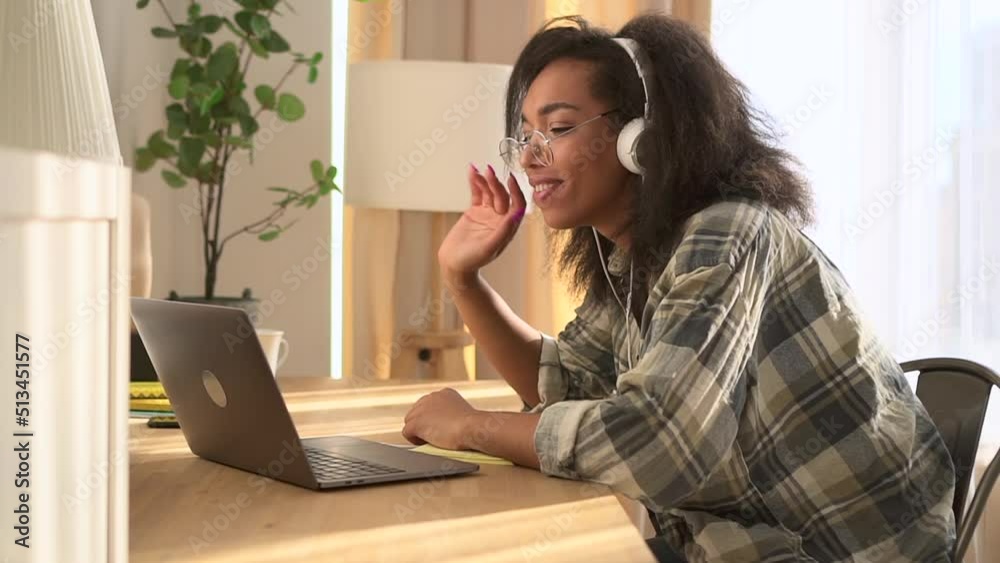 Young woman is waving and talking in front of laptop while sitting at table in home interior spbd. Close view of American African female waves hand and talks online in front of device, works or Young woman is waving and talking in front of laptop while sitting at table in home interior spbd. Close view of American African female waves hand and talks online in front of device, works or