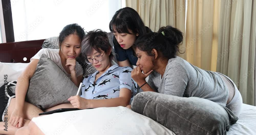 Young Asian girls sleepover pajama party studay red play game on tab on large bed in bedroom with bright light window Young Asian girls sleepover pajama party studay red play game on tab on large bed in bedroom with bright light window