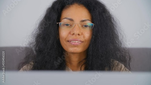 Young black woman in eyeglasses looking in camera with a smile. Portrait of African entrepreneur female working on laptop computer at home. BIPOC person lifestyle video clip photo