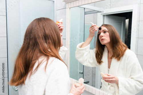 Women's morning routine. A young woman in a white bathrobe puts makeup on her face with a brush