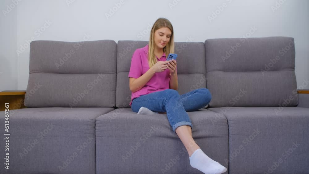 Beautiful Ukrainian female typing message on mobile phone while sitting on couch at home. Cheerful blonde woman communicating online with modern blue smartphone gadget Beautiful Ukrainian female typing message on mobile phone while sitting on couch at home. Cheerful blonde woman communicating online with modern blue smartphone gadget