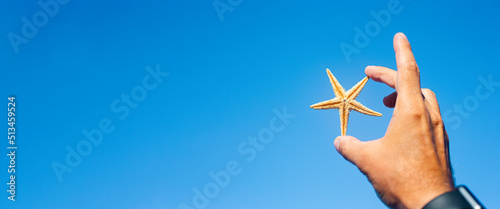Canvas Print Male hand holding a starfish on a blue sky background. Banner.