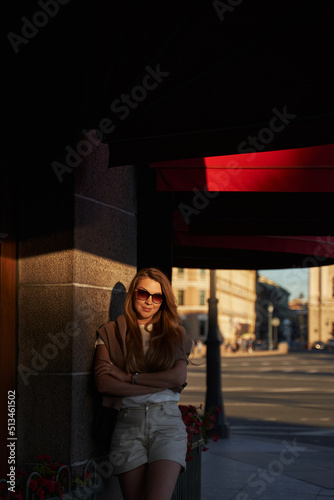 a woman in the sun at the building with high windows and red visors on the windows stands leaning against the wall. city walks in summer, tourism. vacation in the city