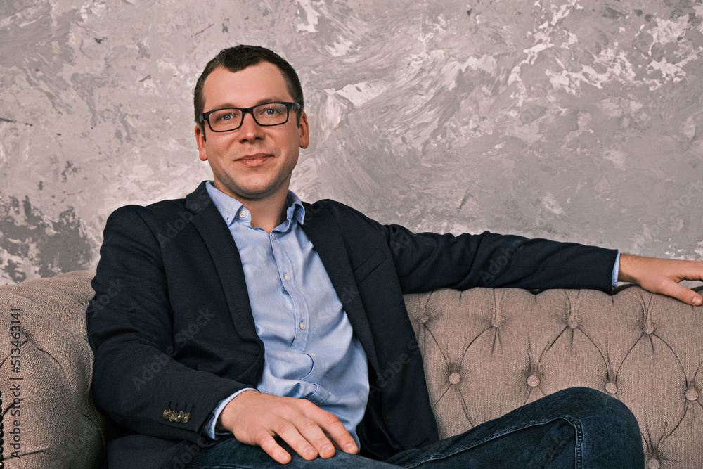 Portrait of young caucasian smiling businessman wearing jacket and glasses sitting on sofa in an open position looking at the camera against background of gray concrete wall. Concept of success Portrait of young caucasian smiling businessman wearing jacket and glasses sitting on sofa in an open position looking at the camera against background of gray concrete wall. Concept of success