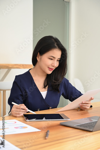 Attractive young business woman holding financial reports and using goal tablet at office desk