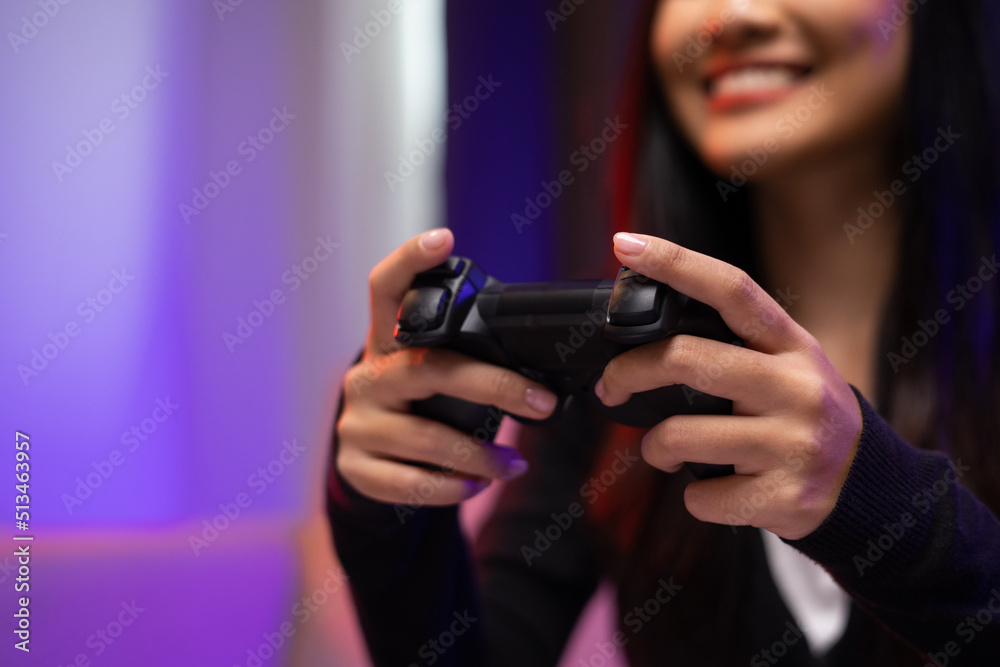 Close up hands Playing video game. Young pretty woman sitting on sofa holding joystick in living room. Female Professional Streamer wearing hoodie playing game online in dark room neon light. Close up hands Playing video game. Young pretty woman sitting on sofa holding joystick in living room. Female Professional Streamer wearing hoodie playing game online in dark room neon light.