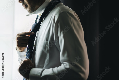 Confident businessman tying or adjust the necktie near window in hotel room in the morning. Handsome man wearing a nice necktie on wedding day. © Chanakon