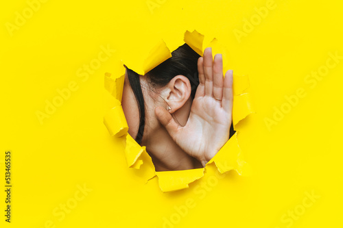 Close-up of a left woman's ear and hand through a torn hole in the paper. Bright yellow background, copy space. The concept of eavesdropping, espionage, gossip and tabloids. photo