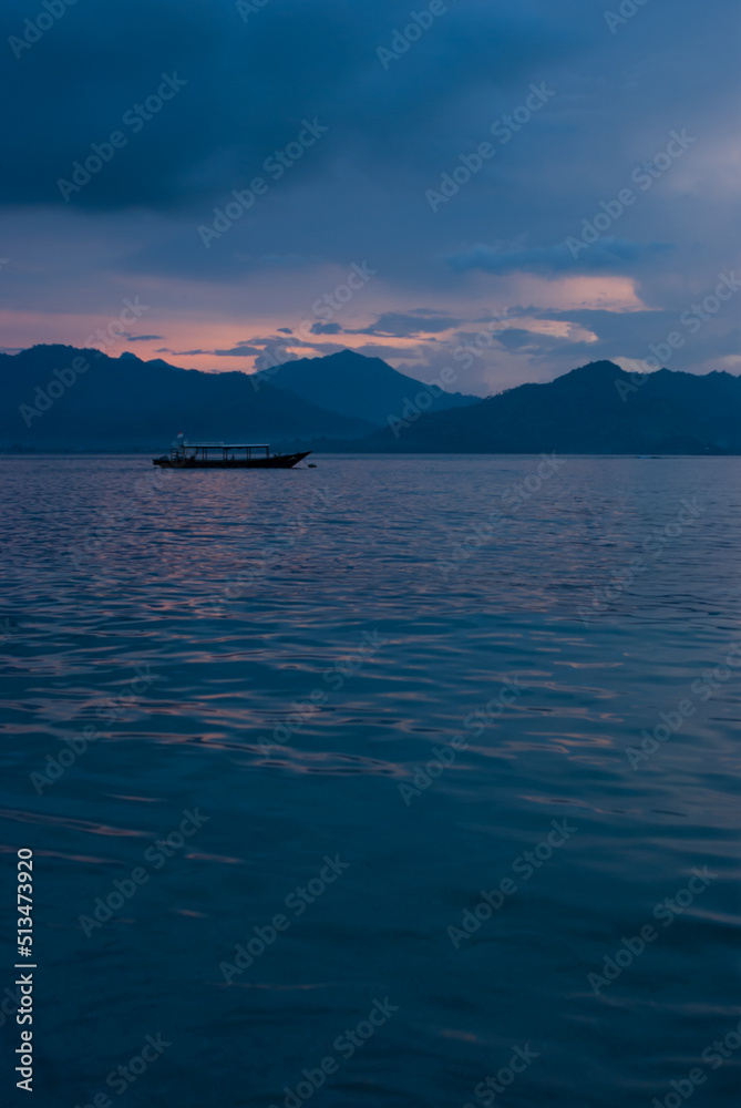 Tender blue evening on ocean with colorful pink heaven, clouds, calm smooth water with glare, blue mountains in haze, floating boat, vertical. Rich saturated indonesian sea landscape, trip in asia.