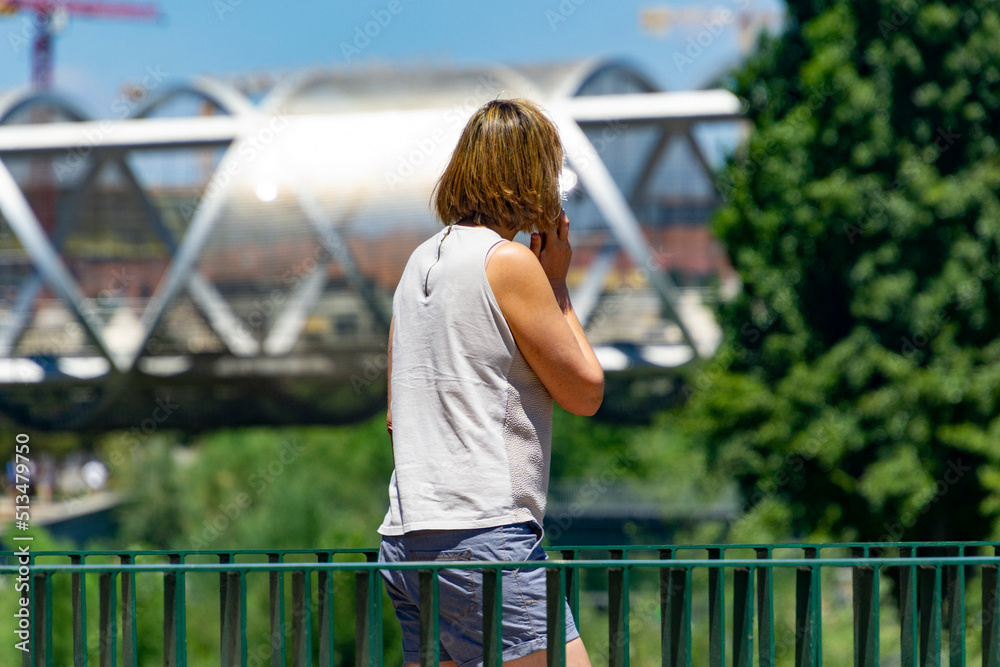 Woman. Person. Middle-aged lady talking on the phone in a park in Madrid. Metal bridge of the Madrid Río park in the background. Clear day, in Spain. Europe. Horizontal photography.