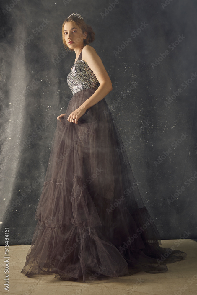 a woman in a long evening dress stands against a dark background, posing relaxed in a smoky room, with a crown on her head