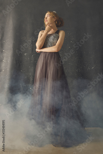 a woman in a long evening dress stands against a dark background, posing relaxed in a smoky room, with a crown on her head © Lina Solntseva 