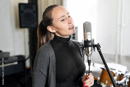 Young Asian or Caucasian brunette woman singing lyric song to microphone alone in professional music studio or concervatory class. Radio host speaking to her listeners in broadcasting radio studio.  photo