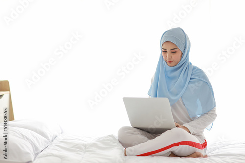 Young asian woman use laptop sitting on bed in bedroom. Beautiful girl in white muslim sleepwear with blue hijab check order or study from notebook before go to bed. Lifestyle and technology concept