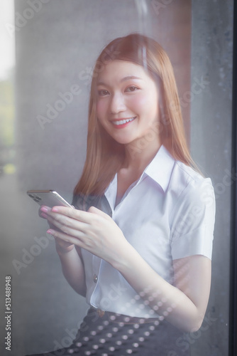 Portrait of an adult Thai student in university uniform. Asian beautiful girl standing near the window happily smiling uses her smartphone to search for educational information.