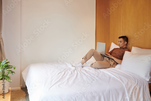Serious Asian young man sitting on bed and coding on laptop