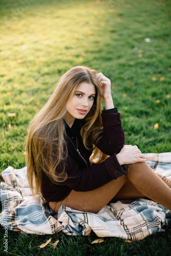Beautiful young girl model with long thick hair sits in an autumn park on a lawn covered with yellow leaves on a plaid blanket and poses. Sunny day, photo in warm tones.