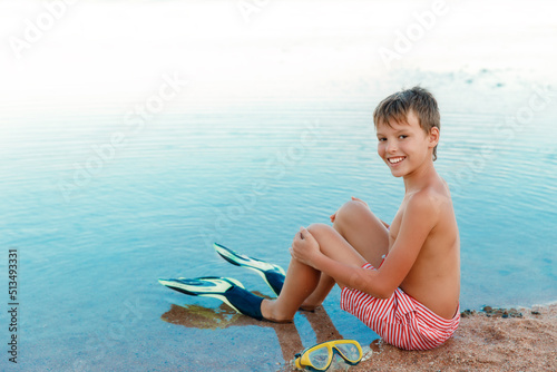 Cute boy at beach enjoyong tropical summer vacation. Kid is sitting on the sand near the sea, wearing swimshorts, flippers and looking at the camera. Summer time on beach.  photo