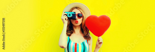 Summer portrait of young woman taking picture on film camera with big red heart shaped balloon wearing straw hat on yellow background