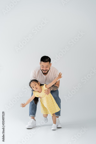 full length of happy asian father playing with daughter in yellow dress standing with outstretched hands on grey.