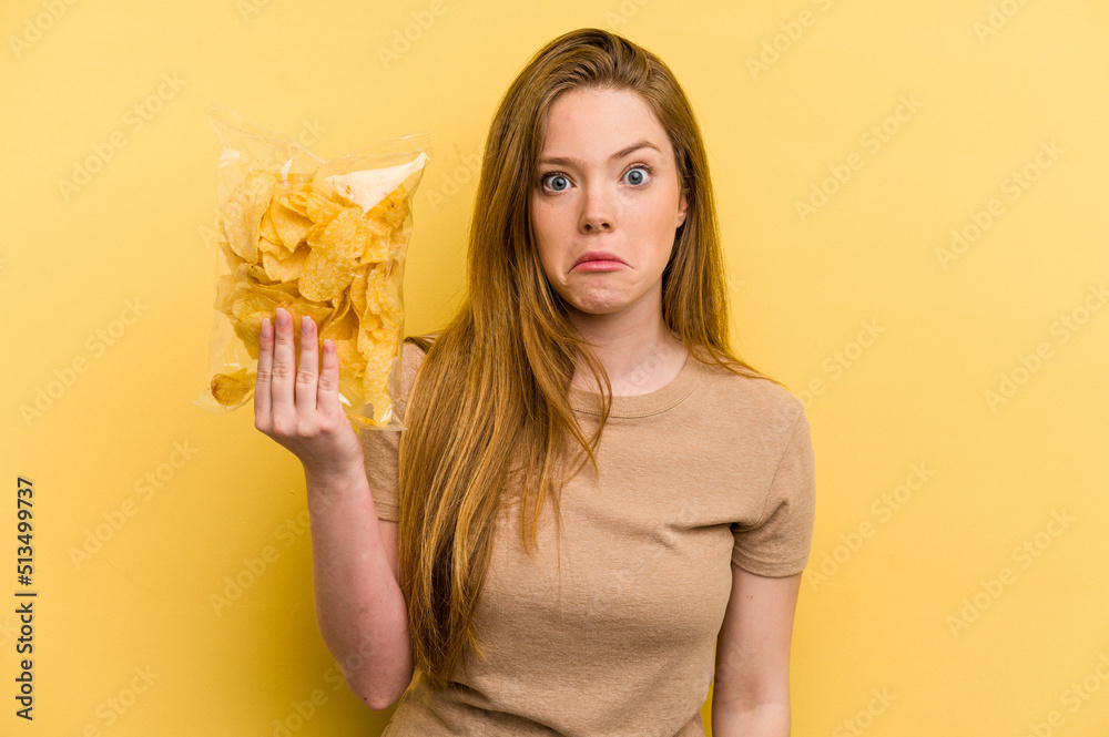 Young caucasian woman holding a bag of chips isolated on yellow background shrugs shoulders and open eyes confused. Young caucasian woman holding a bag of chips isolated on yellow background shrugs shoulders and open eyes confused.