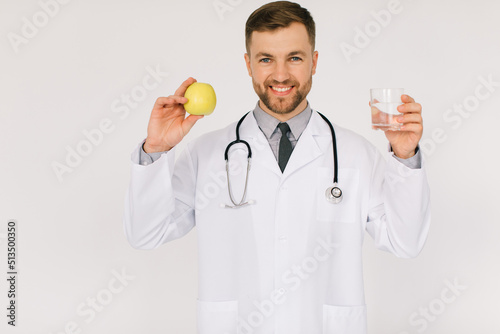 The happy male nutritionist doctor with stethoscope smiling and holding water and apple on white background, diet plan concept photo