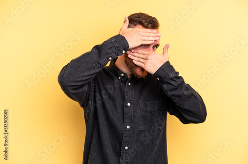 Young caucasian man isolated on yellow background blink at the camera through fingers, embarrassed covering face.