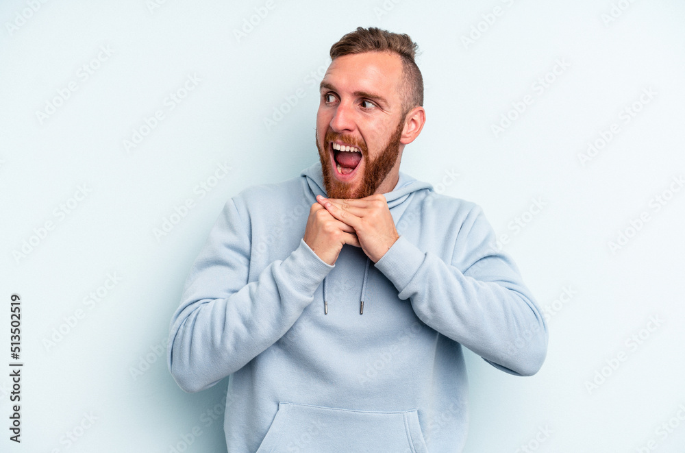 Young caucasian man isolated on blue background keeps hands under chin, is looking happily aside. Young caucasian man isolated on blue background keeps hands under chin, is looking happily aside.