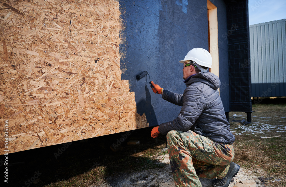 Male painter using paint roller, doing exterior paint work in a black color. Man worker building wooden frame house. Carpentry and construction concept.