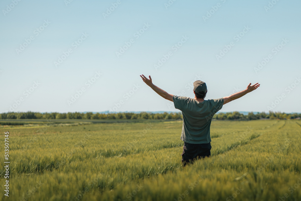 Satisfied successful farmer raising hands in victorious pose in unripe barley crops field on sunny spring day. Rear view of farm worker wearing green t-shirt and trucker hat with arms lifted up. Satisfied successful farmer raising hands in victorious pose in unripe barley crops field on sunny spring day. Rear view of farm worker wearing green t-shirt and trucker hat with arms lifted up.