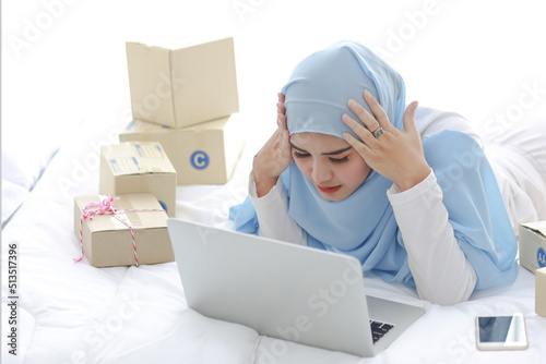 Beautiful and young asian woman in muslim sleepwear with headache emotion lies on bed with computer, mobile phone and online package box delivery. Smart girl with hijab receive bad news and tired © feeling lucky