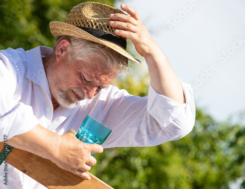 An elderly man sits outside in the great heat. He sweats and drinks mineral water.