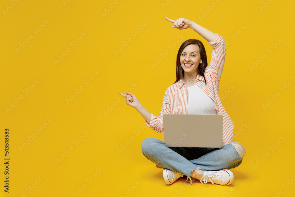 Full body young happy secretary copywriter woman she in striped shirt white t-shirt hold use work on laptop pc computer point index finger aside on workspace area isolated on plain yellow background. Full body young happy secretary copywriter woman she in striped shirt white t-shirt hold use work on laptop pc computer point index finger aside on workspace area isolated on plain yellow background.