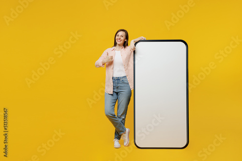 Foto Full body young woman she 30s in striped shirt white t-shirt stand near big huge blank screen mobile cell phone with workspace copy space mockup area show thumb up isolated on plain yellow background