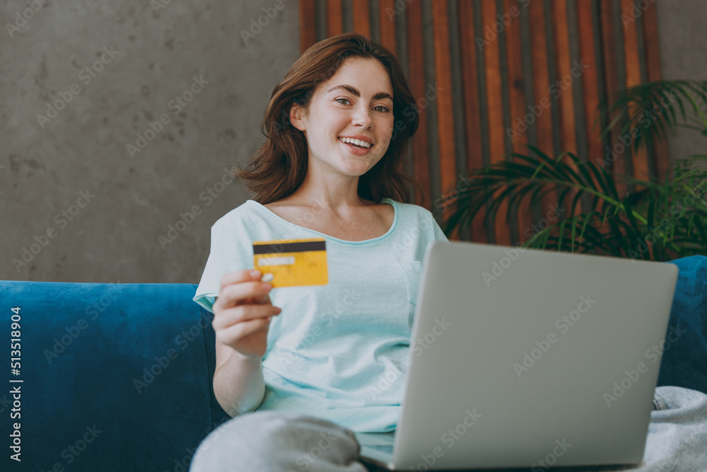 Young happy woman she 20s in mint t-shirt using mobile cell phone hold credit bank card doing online shopping order delivery booking tour sit on blue sofa indoor rest at home in own room apartment Young happy woman she 20s in mint t-shirt using mobile cell phone hold credit bank card doing online shopping order delivery booking tour sit on blue sofa indoor rest at home in own room apartment