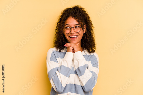 Young Brazilian woman isolated on yellow background keeps hands under chin, is looking happily aside.