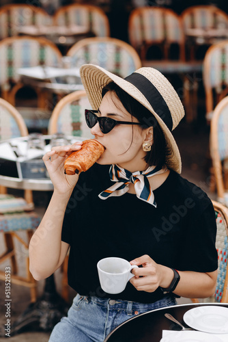 Young beautiful smiling. Carefree woman posing at veranda cafe in the street. Positive model drinking coffee. Enjoying vacation. Eating croissant. In hat and in glasses.