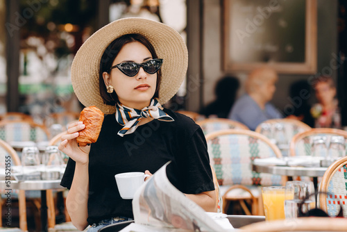 Young beautiful smiling hipster female in trendy summer clothes.Carefree woman posing at veranda cafe in the street. Positive model drinking coffee. Enjoying vacation. In hat and in glasses.