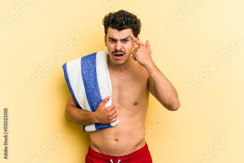 Young caucasian man holding beach towel isolated on yellow background showing a disappointment gesture with forefinger.