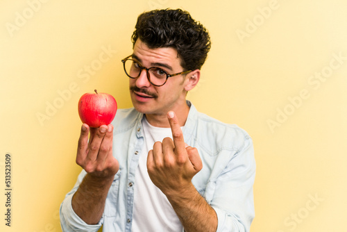 Young caucasian man holding an apple isolated on yellow background pointing with finger at you as if inviting come closer. photo